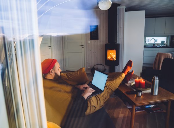 En person med rød lue sitter avslappet i sofaen med en bærbar PC på fanget, mens det brenner i peisen og levende lys står på bordet.