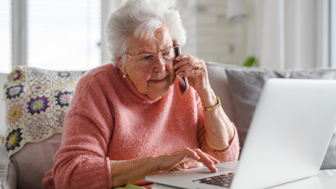 Eldre kvinner sitter i en sofa og snakker i mobiltelefonen samtidig som hun taster på den bærbare datamaskinen sin