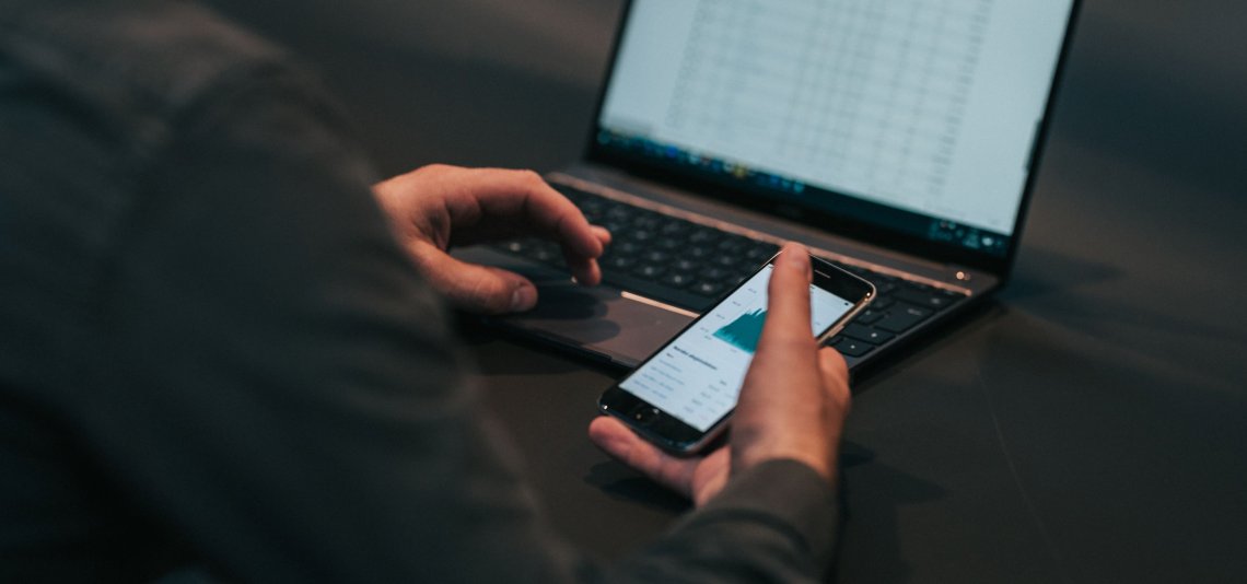 En person sitter ved en åpen laptop og holder en mobiltelefon i hånda.