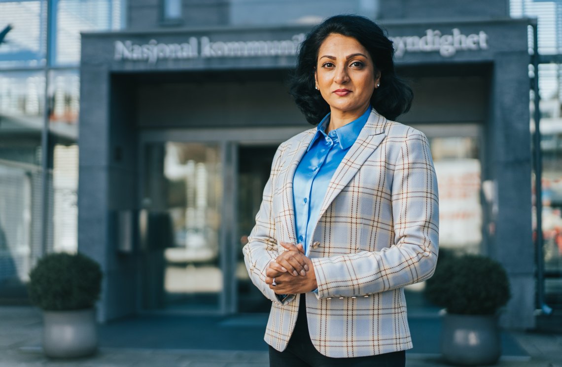 Bildet viser avdelingsdirektør i Nkom, Kamilla Sharma står utenfor inngangsdøra til Nkom. Hun smiler og ser i kamera. ra.