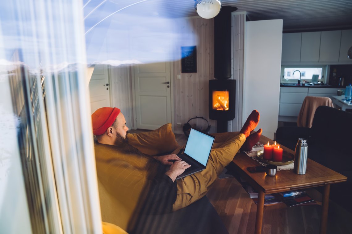 En person med rød lue sitter avslappet i sofaen med en bærbar PC på fanget, mens det brenner i peisen og levende lys står på bordet.