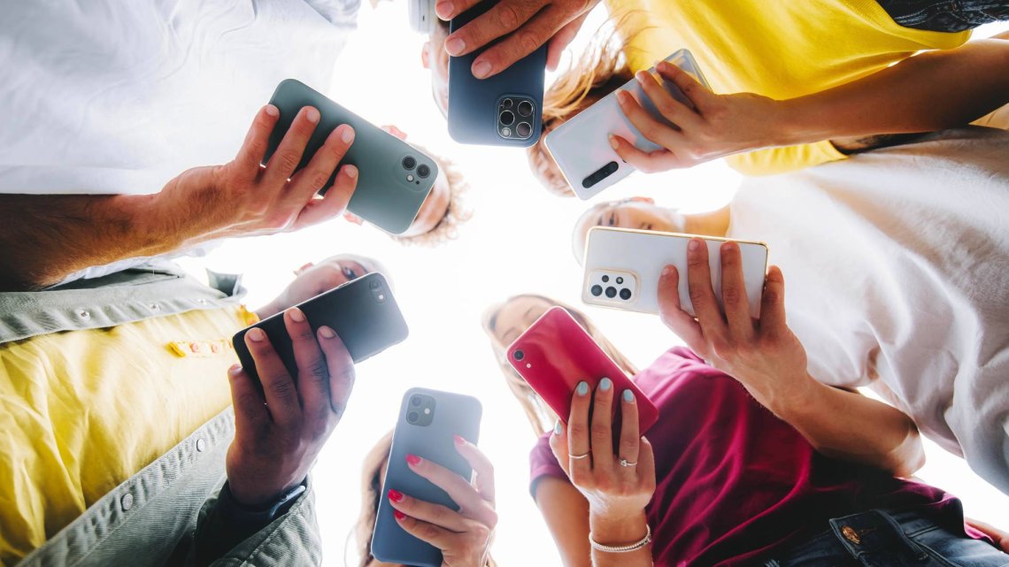 En gruppe mennesker står i en sirkel og holder opp smarttelefoner, sett fra et lavt perspektiv. Hver person ser ned på telefonen sin, og det er en blanding av forskjellige telefonmodeller.