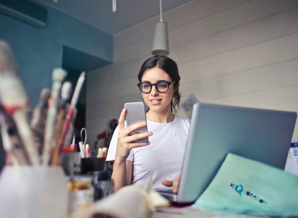 Bilde av jente som sitter foran laptop med mobiltelefon i hånda som hun ser på.