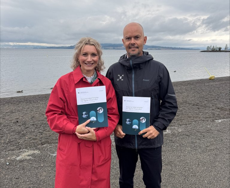 Karianne Tung, digitaliserings‑ og forvaltningsminister, og Nkom‑direktør John‑Eivind Velure, holder hver sin utgave av sikkerhetsplanen for digital infrastruktur ved vannkanten.